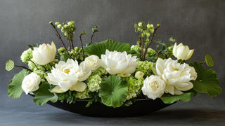 Real touch white florals and green hydrangeas arranged in a low black oval bowl with large leaf accents.