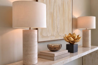 Console table styling with travertine lamps, abstract cream art, a smoked glass bowl on books and a gold lotus sculpture