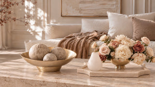 Cream and blush florals beside a gold bowl of decorative spheres on a stone coffee table, styled with a cream sofa and warm neutral accents.
