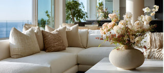 Bright contemporary living room featuring a neutral sectional sofa layered with textured beige pillows, a sculptural stone vase filled with soft blush and ivory florals, and a minimalist modern interior with natural light and ocean views