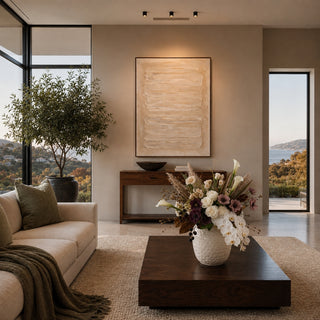 A modern living room with beige walls, a large abstract canvas print hanging above a sofa, a dark wood coffee table with a floral arrangement, and large windows overlooking a scenic outdoor view.