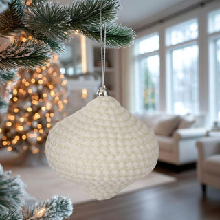 Onion Shaped white and gold beaded Christmas ornament hanging on snow-frosted tree branch in cozy living room – elegant textured holiday décor in neutral tones.