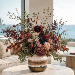 9-inch blush and bronze hand-painted porcelain vase styled with burgundy, mauve, cream, rust, and greenery florals on a round stone table in a light-filled living room.