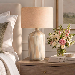 Almeta Lamp with a softly variegated cream, beige, and gray body and natural linen-look drum shade, styled on a warm wood nightstand beside a neutral upholstered bed and floral arrangement.