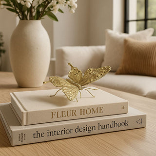 Gold butterfly figurine resting on a stack of neutral books atop a light wood coffee table, with a ceramic vase and soft beige sofa in the background, Third & Main