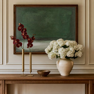 Set of two antiqued brass tall iron vases styled with deep burgundy faux orchid stems on a wood console beneath green abstract artwork.