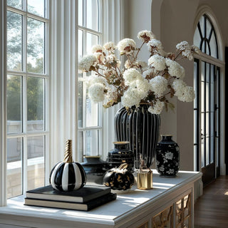 Black and white striped ceramic pumpkin with gold-colored stem styled on window console with black striped vase and white hydrangea arrangement.