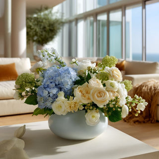 Blue real touch hydrangea stems styled with ivory and pale yellow Duchess roses, cream ranunculus, and green viburnum in modern coastal interior with natural light