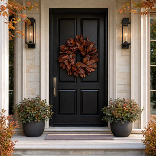 Bourbon Brown magnolia leaf wreath styled on a black front door with dark planters and autumn foliage on a covered entry porch.