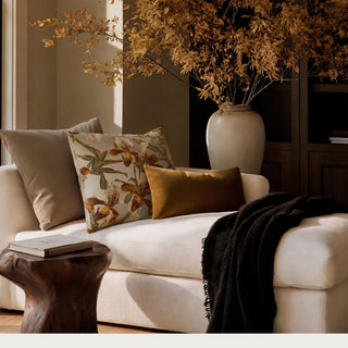 Briarwood Orchid Pillow styled on a neutral sofa with a golden velvet pillow, dark throw blanket, and warm botanical branches in a refined living room.