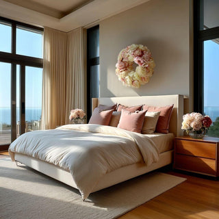 Ultra-realistic 24-inch champagne and blush faux peony wreath styled above a linen upholstered bed in a modern coastal luxury bedroom with floor-to-ceiling windows, warm wood nightstands, and soft natural ocean light.