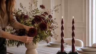 A woman arranging a large floral arrangement in a cream-colored vase on a dining table, with two tall twisted burgundy candles standing beside it in a softly lit room.
