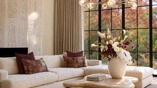 Luxury fall living room with cream sectional, burgundy and rust pillows, marble fireplace, sculptural coffee table, and elegant autumn floral arrangement by large black-framed windows overlooking colorful fall trees.