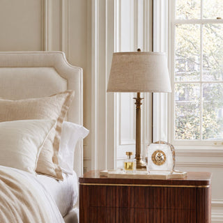 Faceted crystal and gold mantel clock styled on a marble tray atop a warm wood bedroom nightstand beside a linen bed and brass table lamp.