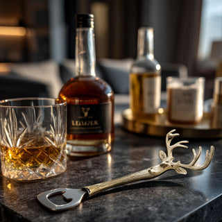Gold and silver stag head bottle opener displayed on a dark marble surface with crystal glassware, whiskey bottle, and candlelight in a modern Santa Monica-style living room, creating a luxurious, cozy holiday entertaining vignette.