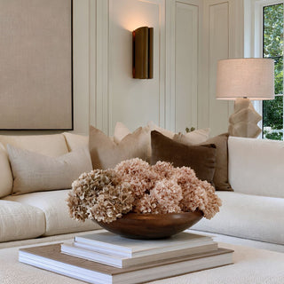A styled living room scene featuring a cream sectional sofa with beige and brown throw pillows, a wooden bowl filled with dried beige hydrangeas on stacked books, and warm ambient lighting from a wall sconce and table lamp.