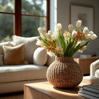Round handwoven seagrass vase filled with white tulips and airy stems, styled on a modern wooden sideboard in a warm, contemporary living room