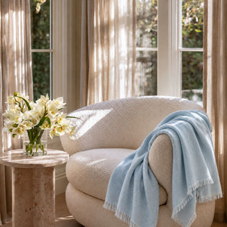 Light blue linen throw over a cream boucle chair beside a vase of white flowers