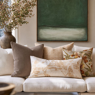 A styled living room scene featuring a cream sofa adorned with multiple decorative throw pillows in neutral, mocha brown linen and floral patterns, with a large framed abstract green painting hanging on the wall behind them.
