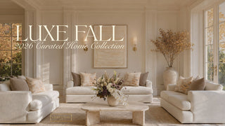 A styled interior scene showing a cream sofa set, matching pillows, a low wooden coffee table with a floral arrangement, and a large vase with dried branches in a bright room. Large overlaid text reads 'LUXE FALL 2026 Curated Home Collection.