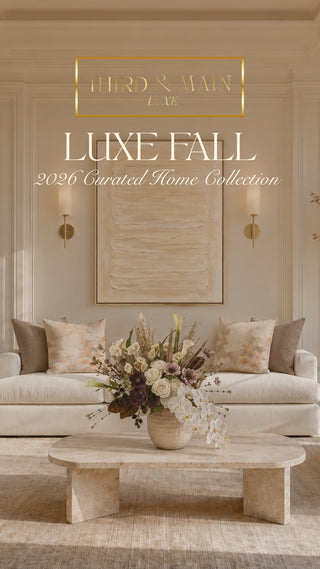 A styled interior scene showing a cream sofa set, matching pillows, a low wooden coffee table with a floral arrangement, and a large vase with dried branches in a bright room. Large overlaid text reads 'LUXE FALL 2026 Curated Home Collection.