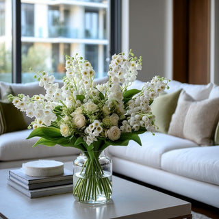 Luxury modern living room styled with a white real touch lily of the valley floral arrangement in a clear glass vase on a contemporary coffee table, featuring lifelike white blooms, lush green leaves, and soft natural light from large windows.
