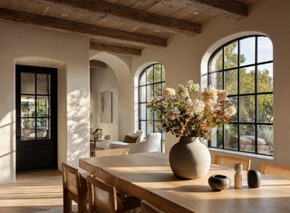 Sunlit Mediterranean-inspired dining room with rustic wood ceiling beams, arched black-framed windows, a natural wood dining table, woven chairs, and a large ceramic vase filled with neutral faux florals.