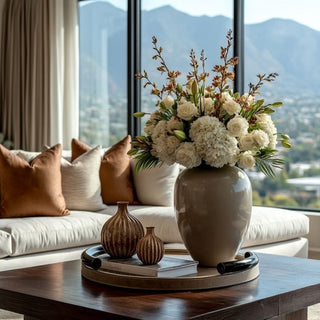 Mink brown leather tray with alloy handles styled on coffee table with large floral arrangement, decorative vases, and books in a modern living room.
