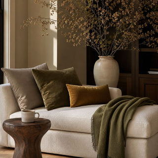 Olive green baby alpaca throw blanket draped over a cream sofa in a warm neutral living room with olive and ochre pillows.