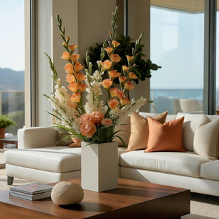 Tall spring-summer floral arrangement featuring orange True Bloom real touch gladiolus stems layered with peach roses, soft white florals, and airy greenery in a matte square ceramic vase on a modern coffee table.