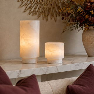 Two cylindrical alabaster candle holders in different heights, glowing with a warm light on a white marble console table in a styled interior setting.