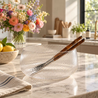 Ribbed clear glass decorative bowl with a wide rounded silhouette and vertical textured detailing, styled with amber-handled serving utensils on a marble kitchen island; available in two sizes.