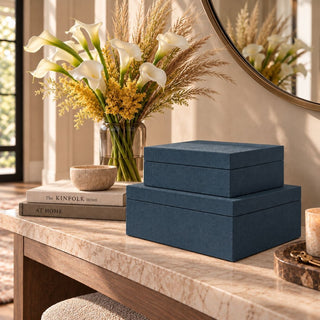 Set of two indigo blue textured decorative boxes in graduated sizes, stacked on a cream stone console table with neutral books and white calla lilies in a warm modern entryway.