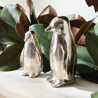 Two silver penguin figurines on a white surface with green leaves in the background perfect for holiday and winter home decor styling 
