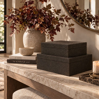 Set of two dark brown textured decorative boxes in graduated sizes, stacked on a cream stone console table with books, a travertine bowl, and autumn branches in a warm modern entryway.