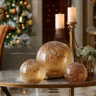Set of three brown and gold speckled glass globes displayed on a round marble table with candles and greenery, warm holiday lighting, and a Christmas tree in the background – handcrafted decorative glass spheres with metallic finish for elegant tabletop or mantel styling in refined holiday décor.