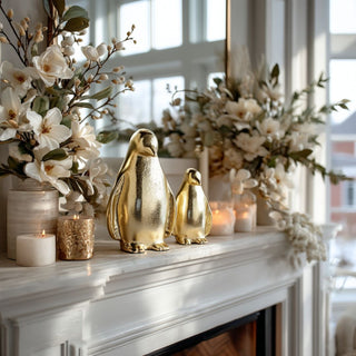Set of two gold aluminum penguin figurines styled with magnolia floral arrangements on a luxury living room fireplace mantel for holiday and winter décor.