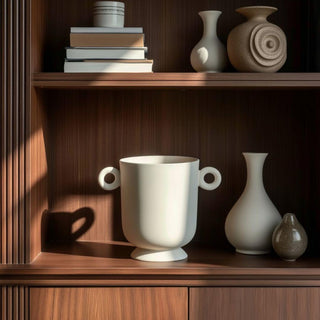 Small white ceramic planter with loop handles styled on a dark fluted wood hutch shelf, surrounded by neutral sculptural ceramics and books in soft morning light