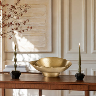 A gold-toned ceramic bowl sits on a wooden console table in a decorated interior, flanked by two black faceted candle holders with green twisted candles. A large framed abstract painting hangs on the wall behind.