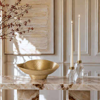 A warm gold textured bowl sits on a marble console table next to two lit white taper candles in faceted clear holders, with a cream paneled wall and framed abstract art in the background.