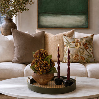 A styled living room scene featuring a cream sectional sofa with textured throw pillows, including a floral patterned pillow, set against a wall with a green abstract painting. In the foreground, a round white marble coffee table holds a dark green tray with a decorative floral arrangement and two dark twisted candles.