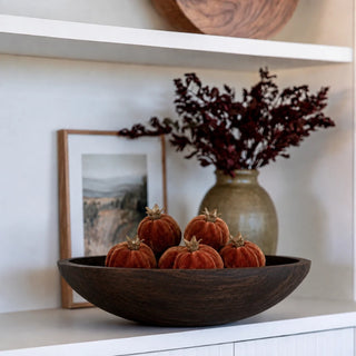 A dark brown wooden bowl filled with mini faux rust/terracotta colored pomegranates, styled on a white shelf next to a ceramic vase with dried burgundy foliage and a framed landscape print.