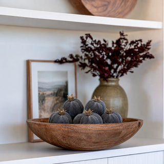 Styled moody blue (dark blue almost purple) faux mini pomegranates styled in a wooden bowl on a shelf with a vase and a floral arrangement in a well lit room.