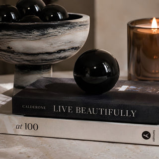 A glossy black decorative glass orb resting on a black hardcover book styled on top of a cream-colored book as well as a  marble bowl filled with black spheres and a lit amber candle are visible in the softly lit background.