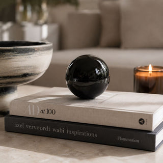 A glossy black crystal glass decorative ball/orb sits on top of two stacked books on a light stone surface, with a softly blurred living-room background including a bowl, candle, and sofa.