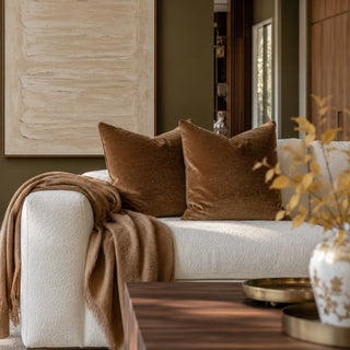 Two brown velvet throw pillows on a white boucle sofa in a styled living room, with a tan throw blanket draped over the arm and a large abstract painting on the wall.