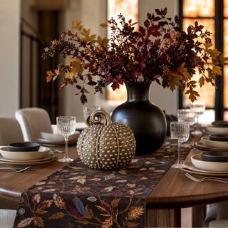 A styled dining table centerpiece featuring a large black vase filled with autumn foliage, a textured metallic pumpkin, and a patterned table runner in a warm indoor setting.