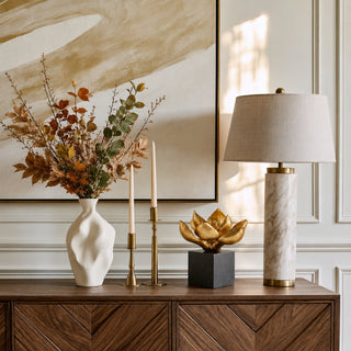 A styled interior scene showing a wooden console table with a white vase of dried leaves, two gold candlesticks, a Edelweiss decorative sculpture, and a table lamp with a marble-look base and beige shade, set against white paneled walls and a large abstract painting.