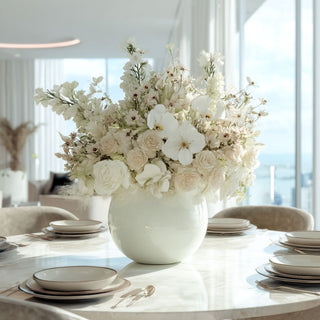 Elegant white faux floral centerpiece with roses, orchids, and real touch  lisianthus in a glossy ceramic vase on a round marble dining table with modern place settings