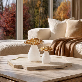 Set of two yellow, white and gold speckled handblown glass white mushrooms styled on a cream and brown colored travertine tray on a coffee table in a modern home with large windows